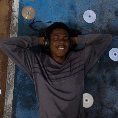 A young man with headphones relaxing on a textured surface surrounded by CDs, enjoying his music. — Jessica Iroh, Pexels
