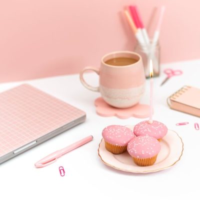 Elegant pink-themed workspace featuring a laptop, cupcakes with candles, and coffee mug. — Jilly Noble, Pexels