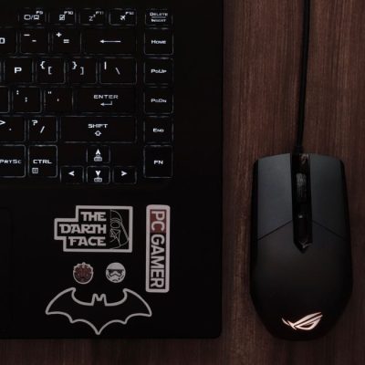 Close-up of a laptop keyboard with a gaming mouse and geek culture stickers on a wooden desk. — Kasra  Askari, Pexels