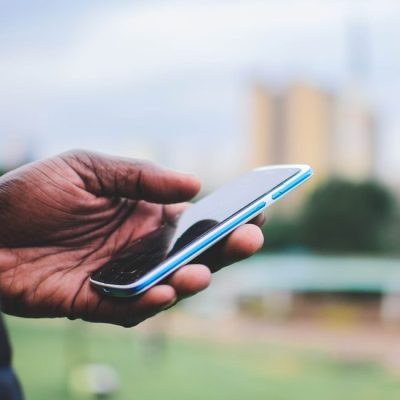 A close-up of a hand holding a smartphone outdoors with blurred urban background. — Mwabonje Ringa, Pexels