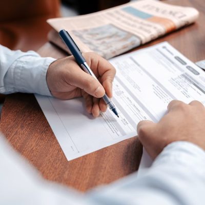 Person reviewing and writing on financial documents with a pen on a wooden desk. — Mohamed hamdi, Pexels